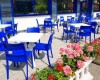 Taras kawiarni z niebieskimi krzesłami i białymi stolikami obok donicy z różowymi kwiatami.