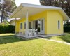 Żółty bungalow z tarasem i plastikowymi krzesłami, otoczony trawnikiem i drzewami.  