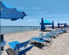 Piaszczysta plaża z niebieskimi parasolami i leżakami nad morzem w słoneczny dzień.