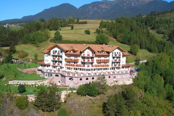 Duży górski hotel otoczony zielenią z górami w tle.