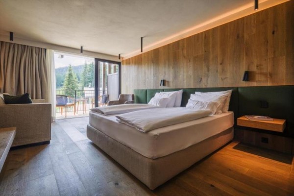 Nowoczesny pokój hotelowy z łóżkiem małżeńskim i widokiem na balkon z lasem w tle.