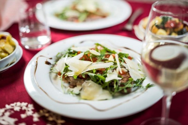 Talerz z carpaccio, rukolą i parmezanem na czerwonym obrusie obok kieliszka białego wina.