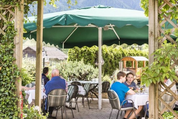Ludzie siedzą przy stołach pod dużym zielonym parasolem w ogrodowej restauracji.