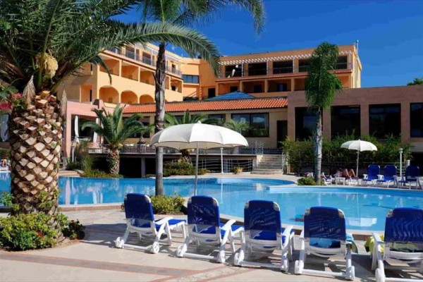 Basen hotelowy otoczony leżakami, palmami i budynkiem w tle.