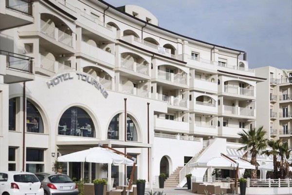 Nowoczesny hotel z jasną elewacją i zewnętrznym tarasem z białymi parasolami.