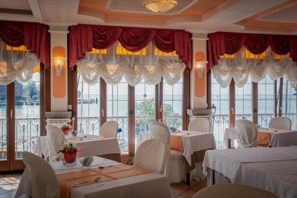 Elegancka restauracja z białymi obrusami, kwiatową dekoracją i widokiem na jezioro przez duże okna.