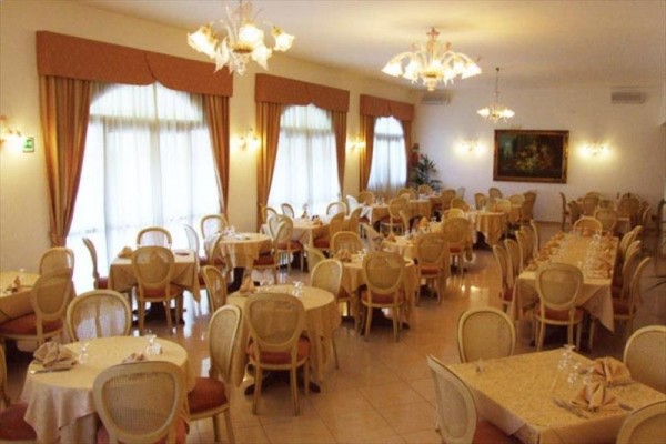 Elegancka restauracja z okrągłymi i prostokątnymi stołami oraz krzesłami, ozdobiona żyrandolami i obrazami.