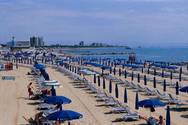 Piaszczysta plaża z niebieskimi parasolami i leżakami nad morzem w słoneczny dzień.