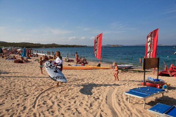 Słoneczna plaża pełna ludzi, z czerwonymi żaglami windsurfingowymi i kobietą niosącą deskę surfingową.