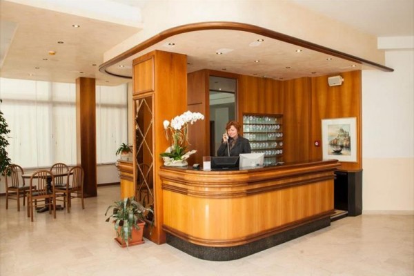 Recepcja hotelowa z drewnianym blatem i kwiatami, przy której stoi recepcjonistka.