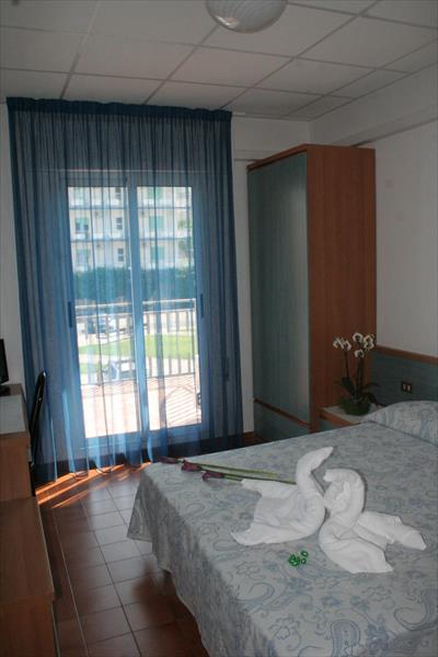 Pokój hotelowy z łóżkiem małżeńskim, zasłoną i kwiatem na stoliku.