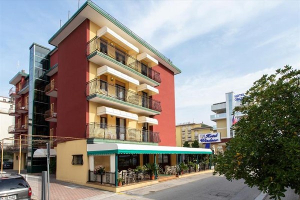 Czteropiętrowy hotel z werandą i balkonami, otoczony zielenią i sąsiednimi budynkami.