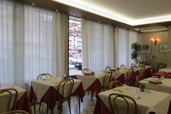 Restauracja z nakrytymi stołami, czerwonymi serwetkami i drewnianymi krzesłami przy dużych oknach.
