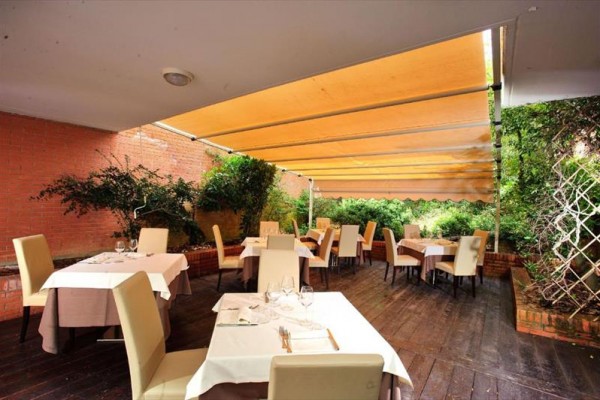 Restauracja z zewnętrznym tarasem i stolikami pokrytymi obrusami, otoczona zielenią.