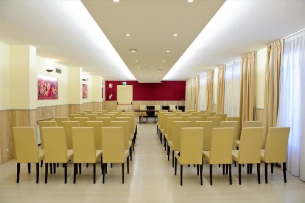 Sala konferencyjna z rzędami beżowych krzeseł i pustą tablicą z przodu.