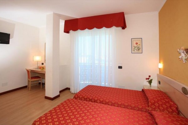 Dwa pojedyncze łóżka z czerwonymi narzutami w pokoju hotelowym; w tle biurko z krzesłem i balkon.