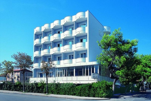 Czteropiętrowy biały hotel z balkonami, otoczony drzewami i krzewami, pod błękitnym niebem.