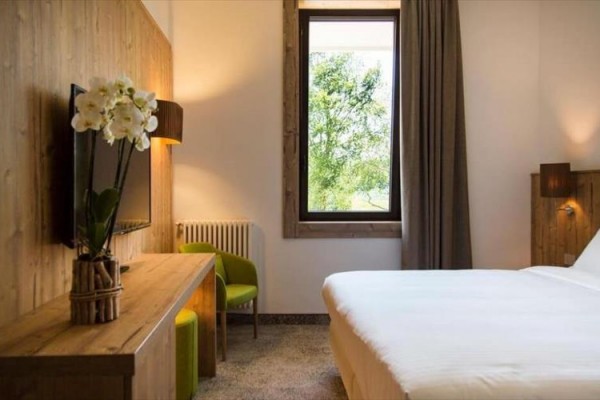 Nowoczesny pokój hotelowy z białym łóżkiem, zielonymi fotelami i kwiatem na drewnianym stole przy oknie.