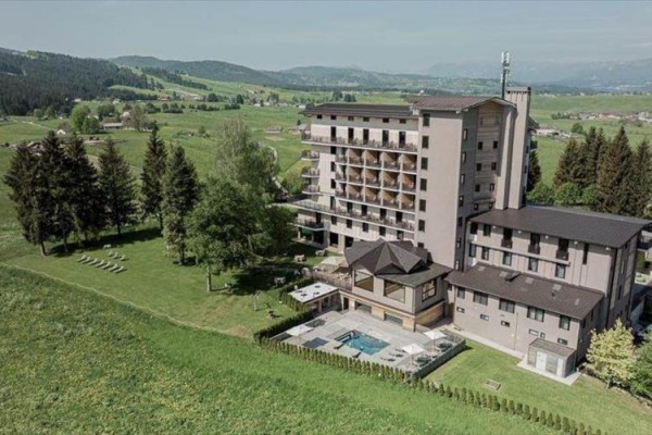 Duży kompleks hotelowy otoczony zielonymi polami i drzewami w górskim krajobrazie.