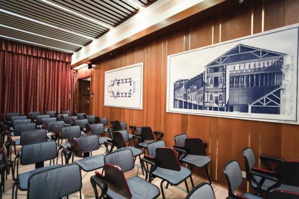 Sala z rzędami pustych krzeseł i planami architektonicznymi na ścianach.