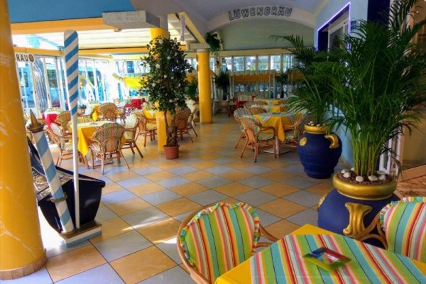Słoneczna restauracja z kolorowymi stolikami i krzesłami, otoczona roślinami.