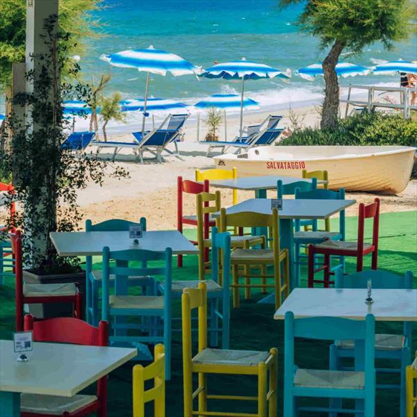 Kolorowe krzesła i stoły na tarasie z widokiem na plażę i morze z niebieskimi parasolami.