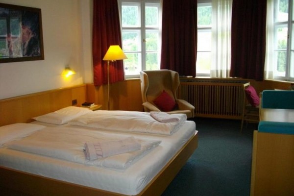 Pokój hotelowy z łóżkiem małżeńskim, fotelem i oknami z widokiem na zieleń.