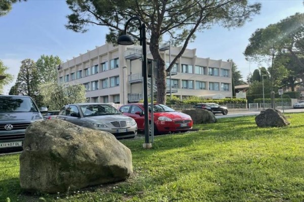 Budynek znajduje się za parkingiem z samochodami, otoczony drzewami i trawnikiem z kamieniami.