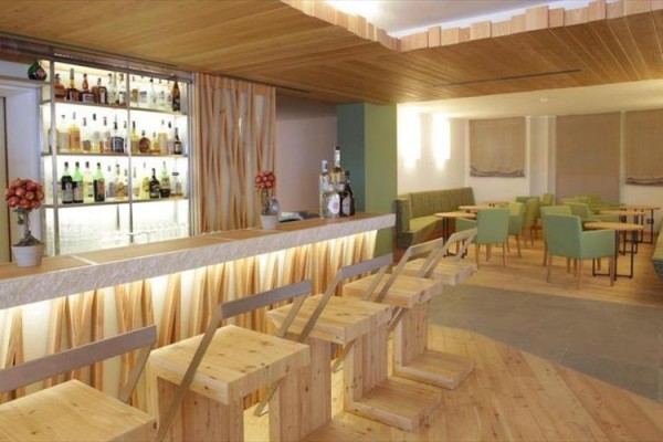 Nowoczesny bar z drewnianymi krzesłami i ławami, podświetlony bar z butelkami alkoholu na półkach w tle.
