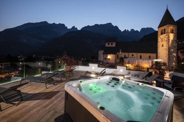 Taras na dachu z jacuzzi, leżakami i widokiem na góry i kościół o zmierzchu.
