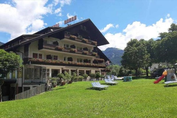 Hotel z dekoracjami na balkonach i leżakami na trawniku, w tle góry.