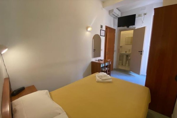 Prosty pokój hotelowy z łóżkiem małżeńskim, toaletą i telewizorem na ścianie.