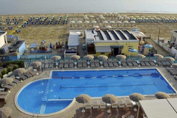 Basen pływacki z leżakami i parasolami przy plaży z morzem w tle.