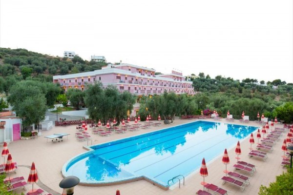 Basen z leżakami i parasolami przed różowym budynkiem hotelu otoczonym zielenią.