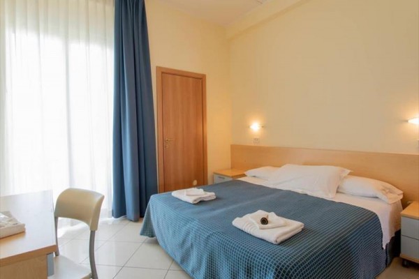 Jasny pokój hotelowy z łóżkiem podwójnym, niebieską pościelą, dwoma stolikami nocnymi i zasłonami.