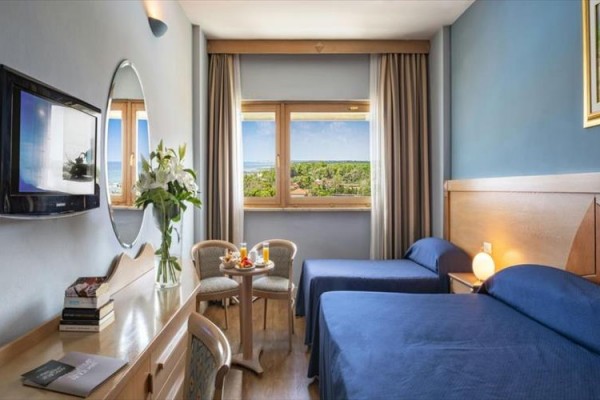 Pokój hotelowy z dwoma pojedynczymi łóżkami, stolikiem z krzesłami i telewizorem na ścianie.