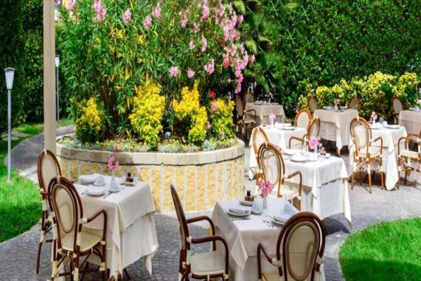 Ogrodowa restauracja z okrągłymi stołami pokrytymi białymi obrusami i kwietnikami z krzewami i kwiatami.