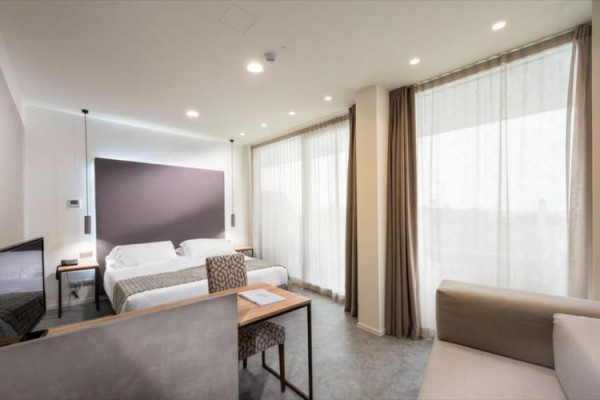 Nowoczesny pokój hotelowy z podwójnym łóżkiem, telewizorem i dużymi oknami z białymi zasłonami.