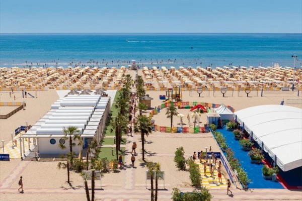 Piaszczysta plaża z rzędami parasoli nad morzem, otoczona letnimi obiektami i zielenią.