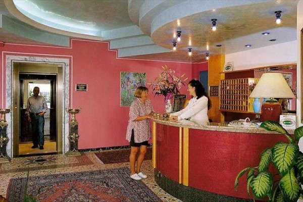 Recepcja hotelowa z kobietą stojącą przy ladzie i osobą wychodzącą z windy.