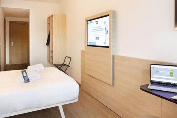 Nowoczesny pokój hotelowy z łóżkiem, dużym ekranem na ścianie i laptopem na stole.