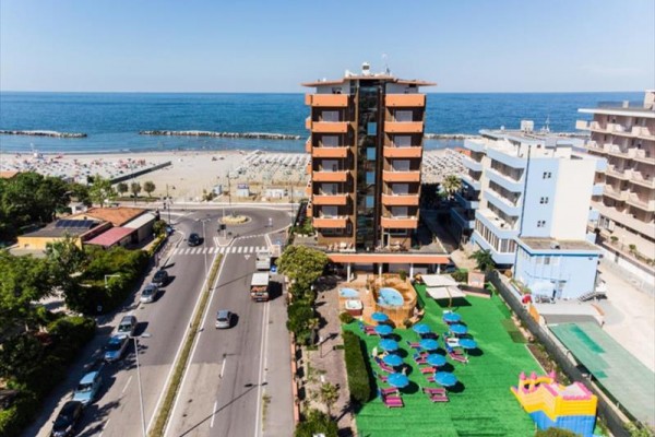 Wysoki budynek nad morzem z trawnikiem i leżakami plażowymi na pierwszym planie.