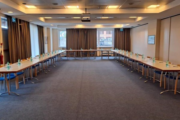 Sala konferencyjna z układem stołów w kształcie litery U i butelkami wody na każdym stole.