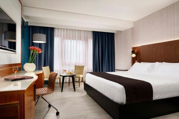 Nowoczesny pokój hotelowy z łóżkiem małżeńskim, biurkiem, krzesłem i małym stolikiem z kieliszkami wina.