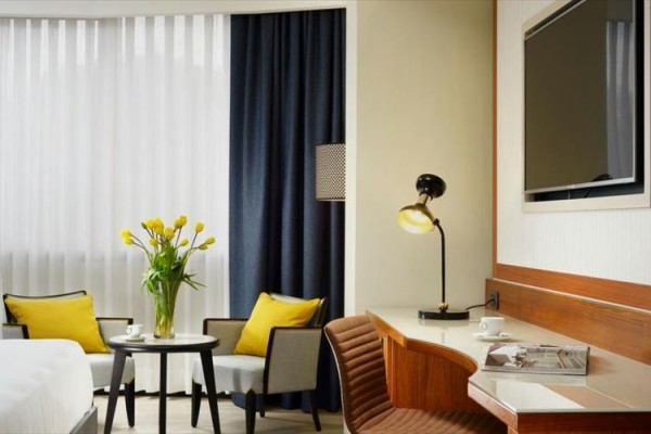 Nowoczesny pokój hotelowy z fotelami, stołem i żółtymi poduszkami przy oknie z ciemną zasłoną.