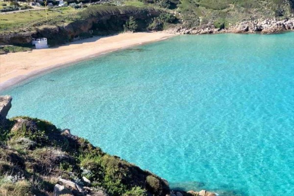Spokojna piaszczysta plaża z czystym, błękitnym morzem otoczona skałami i zielonymi wzgórzami.
