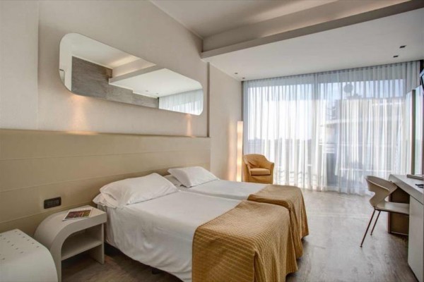 Nowocześnie urządzony pokój hotelowy z dwoma łóżkami, krzesłem i biurkiem obok dużego okna z białymi zasłonami.