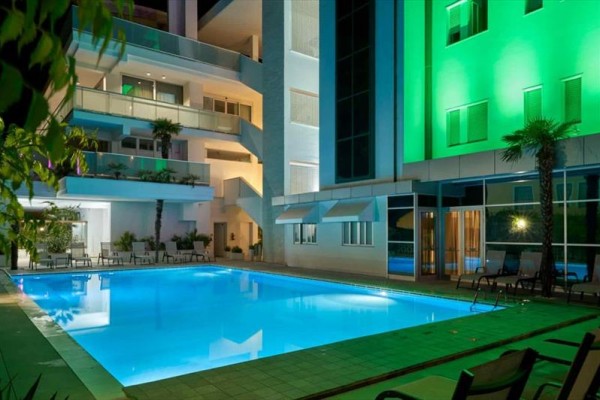 Nocny widok na oświetlony basen hotelowy z leżakami i tropikalnymi roślinami.