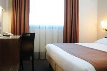 Prosto urządzony pokój hotelowy z łóżkiem, biurkiem i oknem z brązowymi zasłonami.