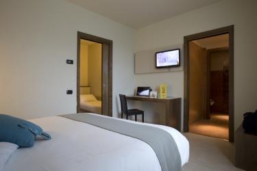 Nowoczesny pokój hotelowy z łóżkiem, biurkiem, dwojgiem drzwi i telewizorem na ścianie.